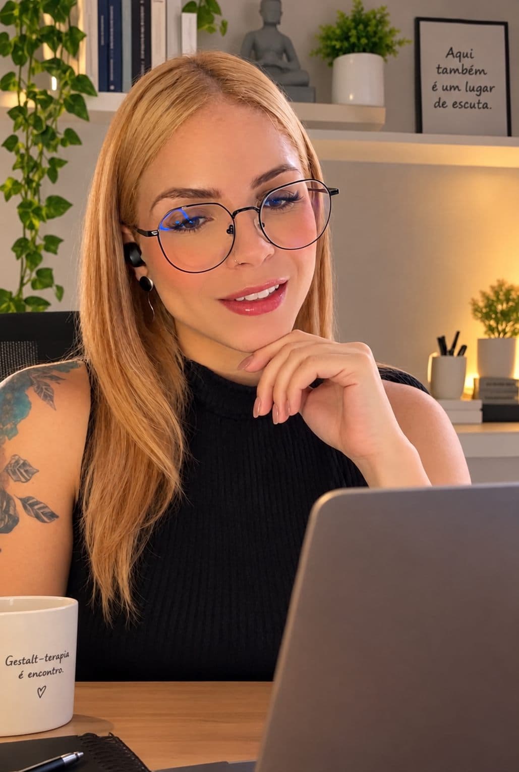 Naila Rayanne sorrindo durante um atendimento online, sentada diante do notebook em seu consultório com luz quente e plantas ao fundo