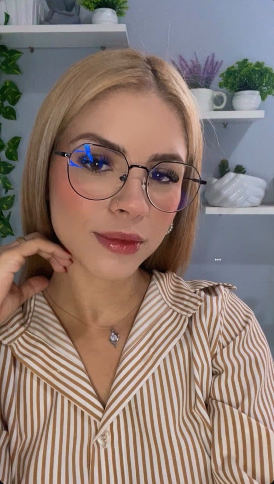 Retrato de Naila Rayanne usando óculos e camisa listrada caramelo, com prateleiras e plantas ao fundo